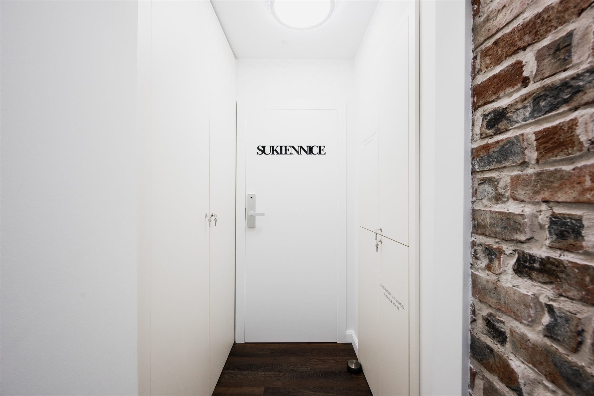 Biały korytarz z drzwiami podpisanymi „SUKIENNICE” i ceglaną ścianą