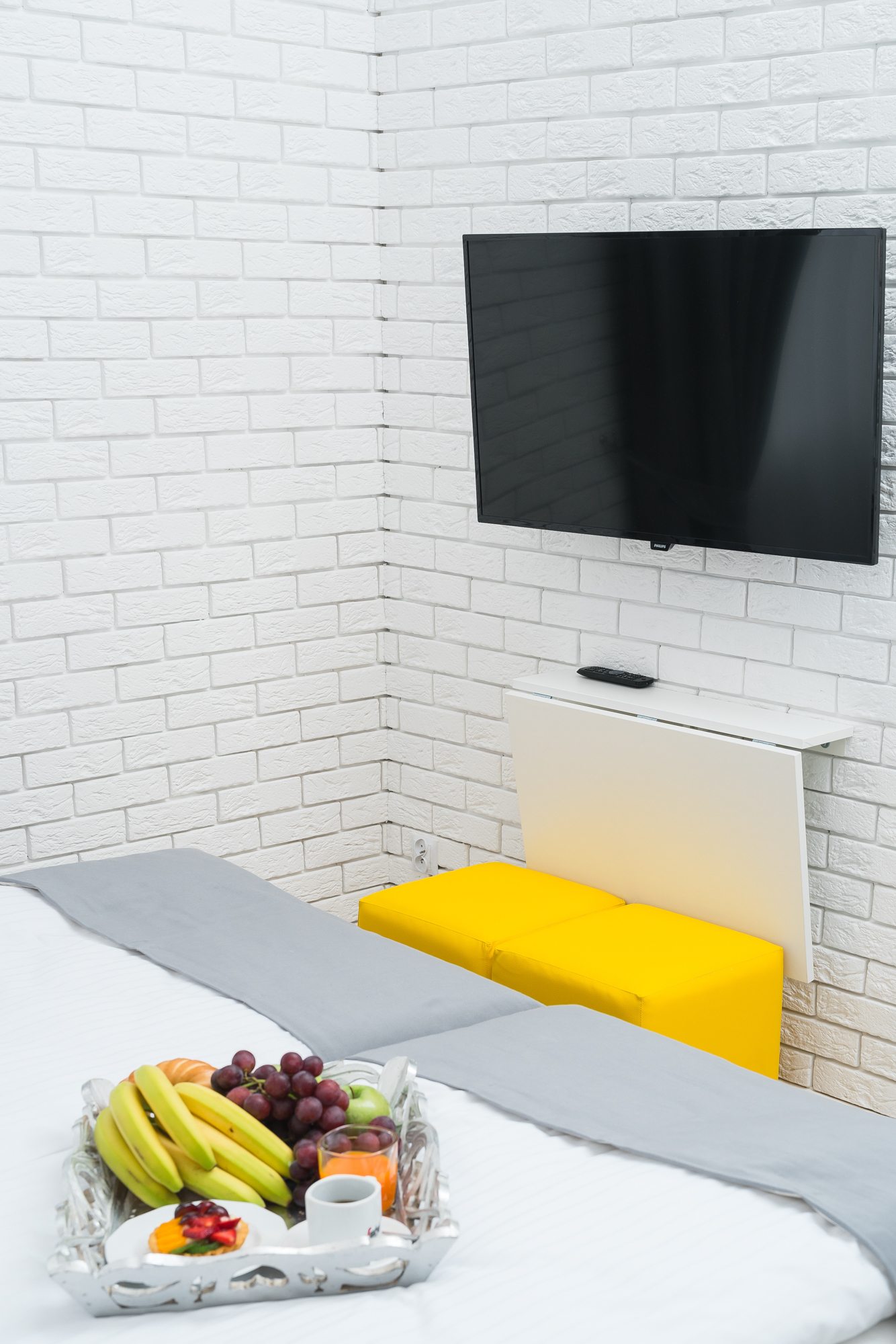 Łóżko z szarym prześcieradłem, telewizor na białej ścianie, owocowy kosz