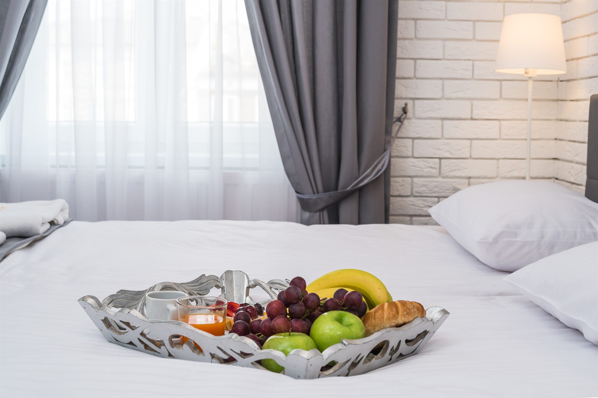 Na łóżku znajduje się srebrna taca z owocami i sokiem.