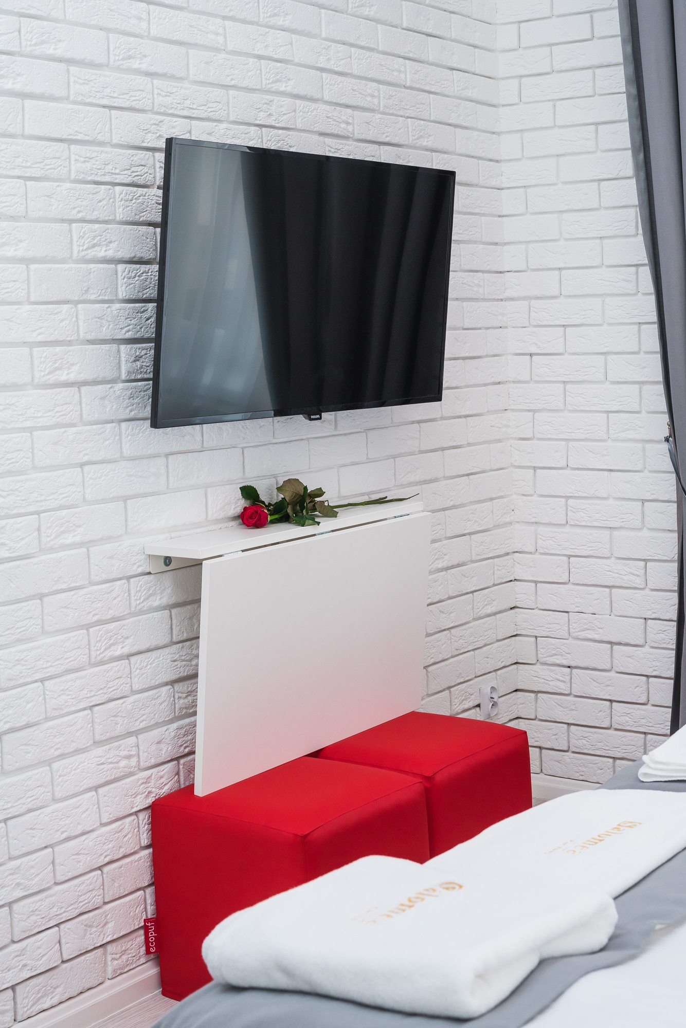Telewizor na białej, ceglanej ścianie obok czerwonych siedzisk i róży
