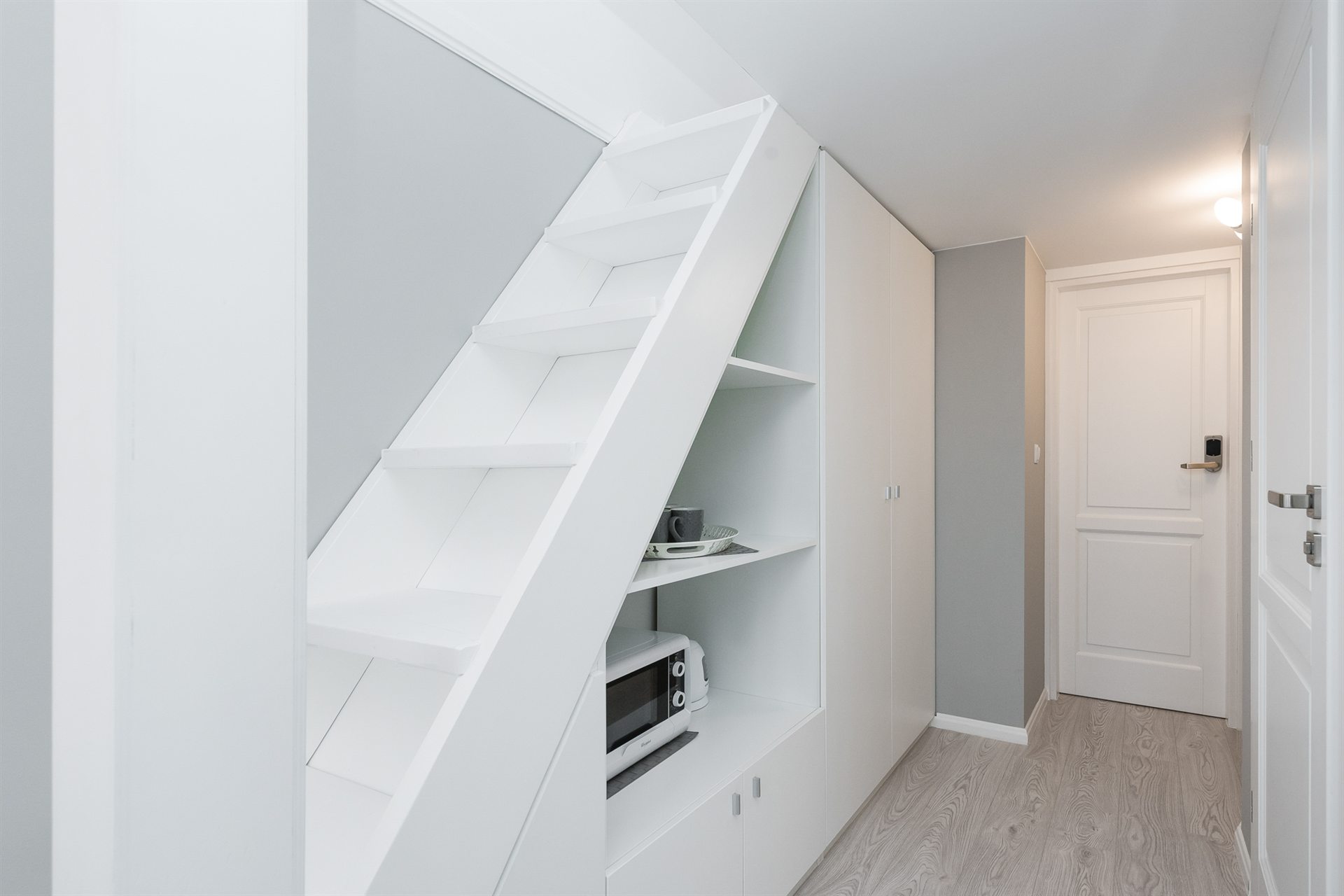 Wnętrze z białymi schodami prowadzącymi do górnej części, obok szafy z półkami. Na dolnej półce zawartość: mikrofalówka, filiżanki i talerz. Szare ściany.