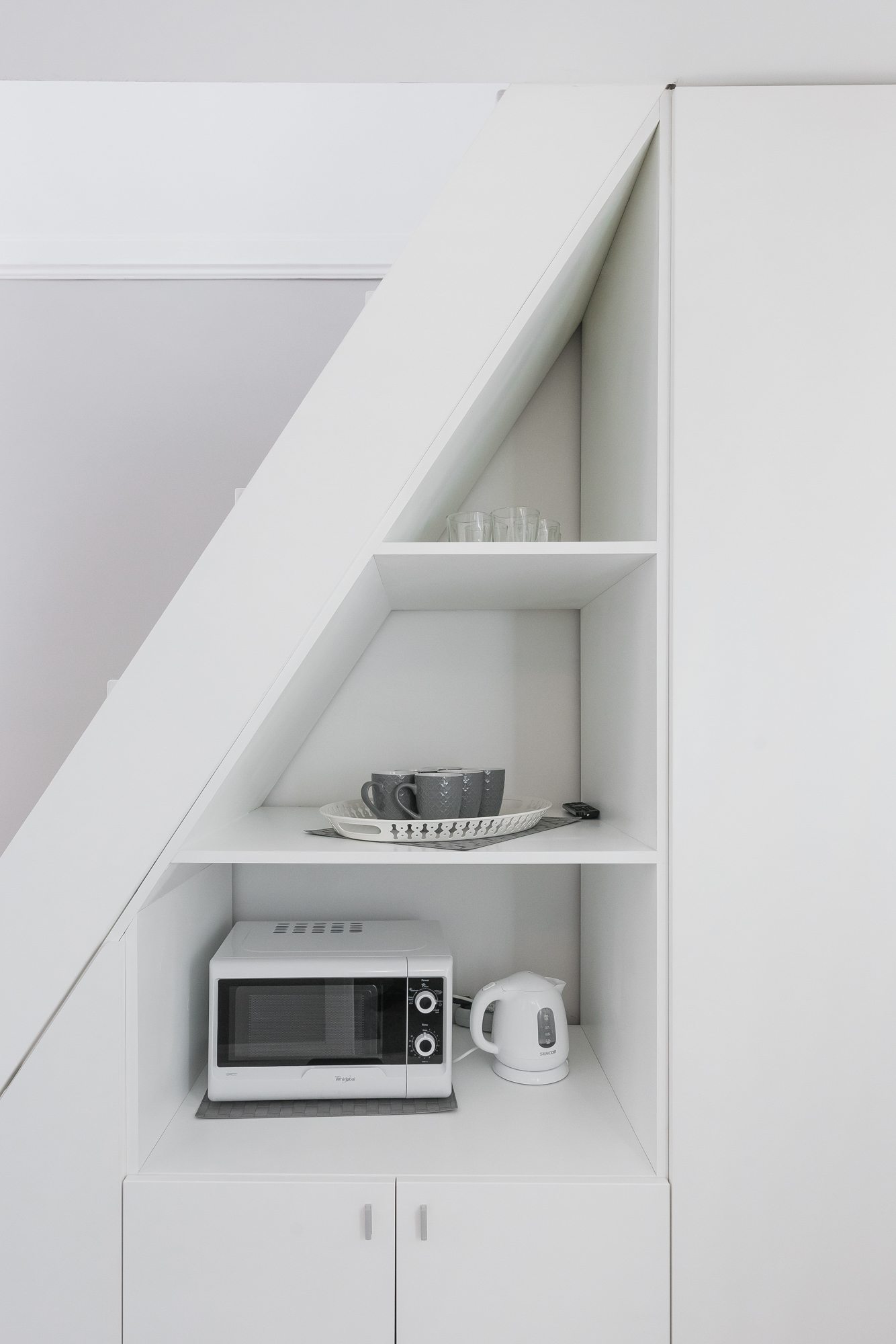 Biała kuchnia z mikrofalówką, czajnikiem i filiżankami na półce